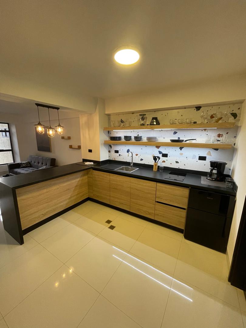 The modern kitchenette features sleek cabinetry with a combination of wood and dark finishes. A wall-mounted shelf displays dishware, while a central sink and a coffee maker are visible on the countertop. Soft lighting highlights the contemporary design and clean lines.