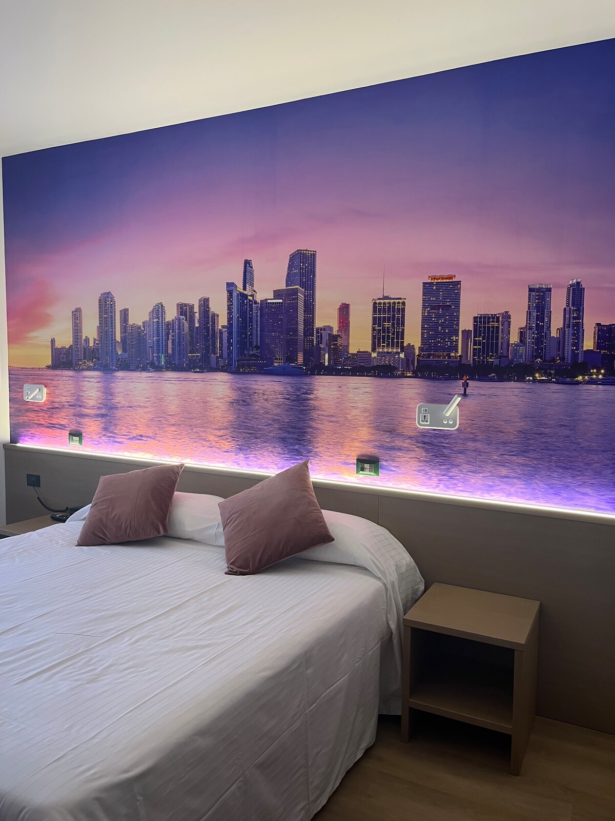 A spacious sleeping area is highlighted by a large mural depicting a city skyline at sunset. Soft pink pillows are placed on the bed, and ambient lighting enhances the modern design. A bedside table complements the contemporary decor.