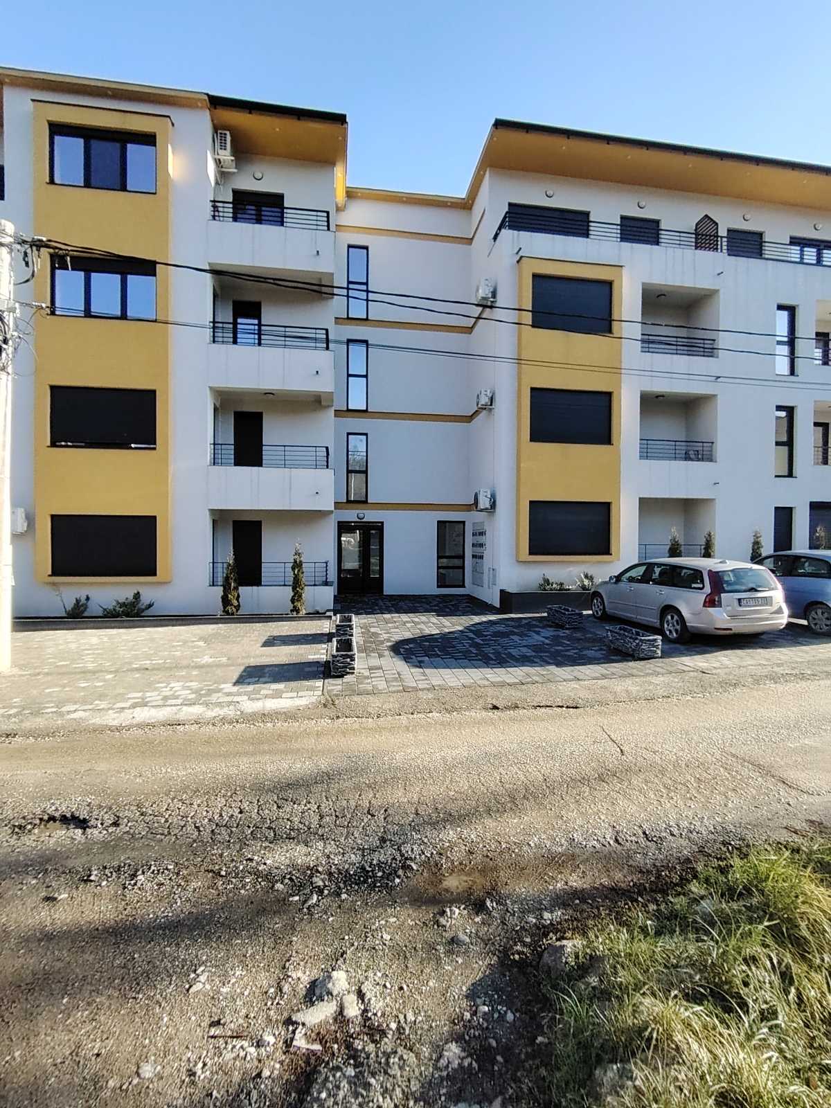 The exterior of a modern apartment building is shown, featuring three floors with balconies and large windows. A neatly arranged parking area is visible in front, bordered by small shrubs. The building's façade is painted in light colors, complemented by dark window frames.