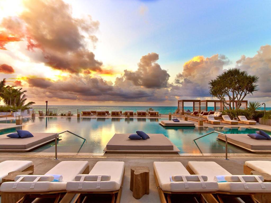 A serene pool area is visible, featuring lounge chairs positioned around the water's edge. The pool's surface reflects the vibrant colors of the sunset, while palm trees and a lounge area are seen in the background. Soft cushions are placed on the chairs for added comfort.