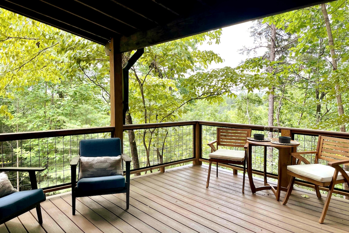 A spacious private deck is surrounded by lush green trees, providing a peaceful outdoor retreat. Comfortable seating includes two accent chairs and a small dining table, perfect for sipping coffee or enjoying a meal while taking in the natural landscape.