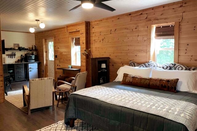 A spacious studio features wooden walls, providing a warm atmosphere. A king bed, adorned with decorative pillows, takes center stage, complemented by cozy seating with a side table. The kitchenette is visible in the background, showcasing modern appliances against a rustic setting.