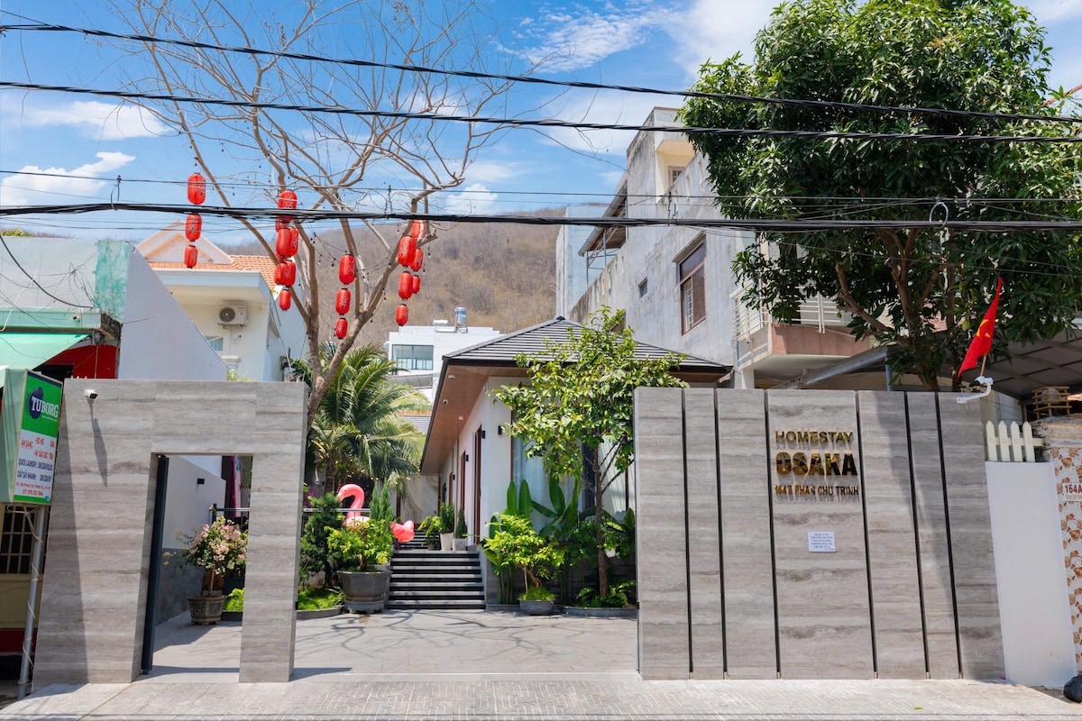 The entrance to Osaka Villa is framed by decorative greenery and stone walls, featuring a prominently displayed sign. Red lanterns hang overhead, and steps lead towards the entrance, creating an inviting pathway. The background reveals adjacent buildings and landscaped areas, contributing to a welcoming environment.