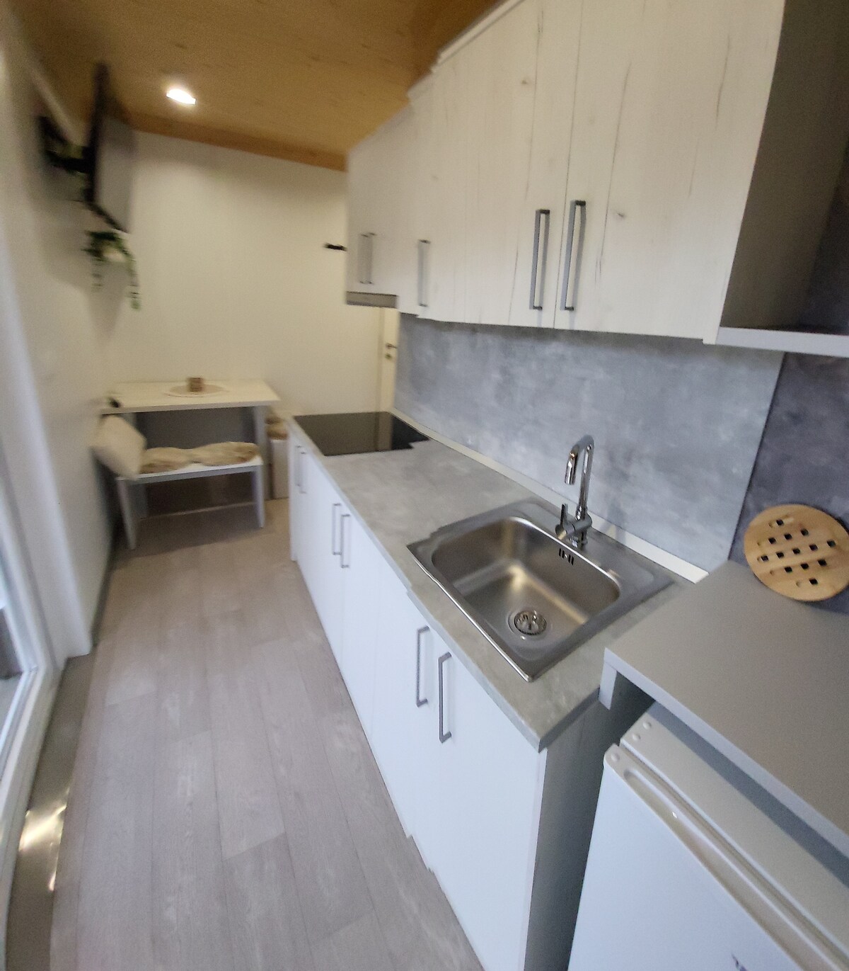A compact kitchen area is presented with white cabinetry and a sleek countertop. A stainless steel sink is visible, accompanied by modern fixtures. Light-colored wood finishes are featured on the ceiling, and a small table is situated at the far end, enhancing functionality.