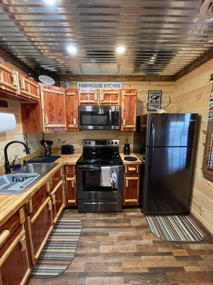 A compact kitchen with wooden cabinetry and counters displays stainless steel appliances, including an oven and refrigerator. A farmhouse-style sink is positioned next to a microwave, while decorative signage hangs above. Colorful mats line the floor, adding a touch of comfort to the space.