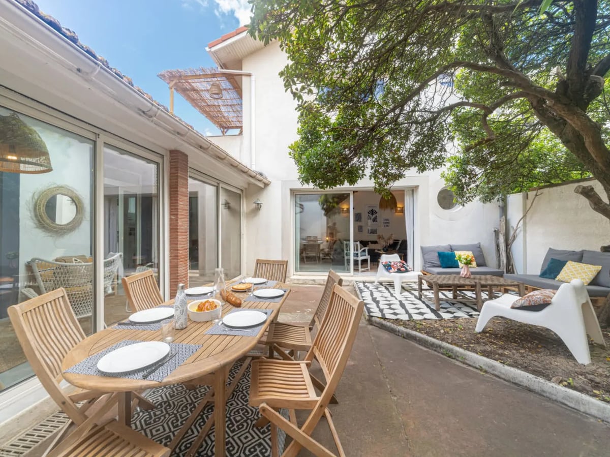 An outdoor patio area features a large wooden dining table surrounded by matching chairs. A cozy seating arrangement is situated nearby, accompanied by a tree providing shade. Decorative elements are present, adding character to the space, while large glass doors connect to the interior.