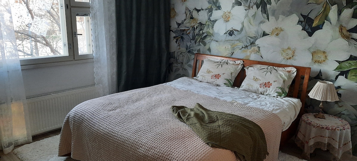 A tranquil bedroom features a large bed adorned with floral bedding and a soft throw draped over one side. An accent wall with a floral mural adds color, while natural light filters through two windows covered with sheer curtains. A small bedside table holds a lamp.