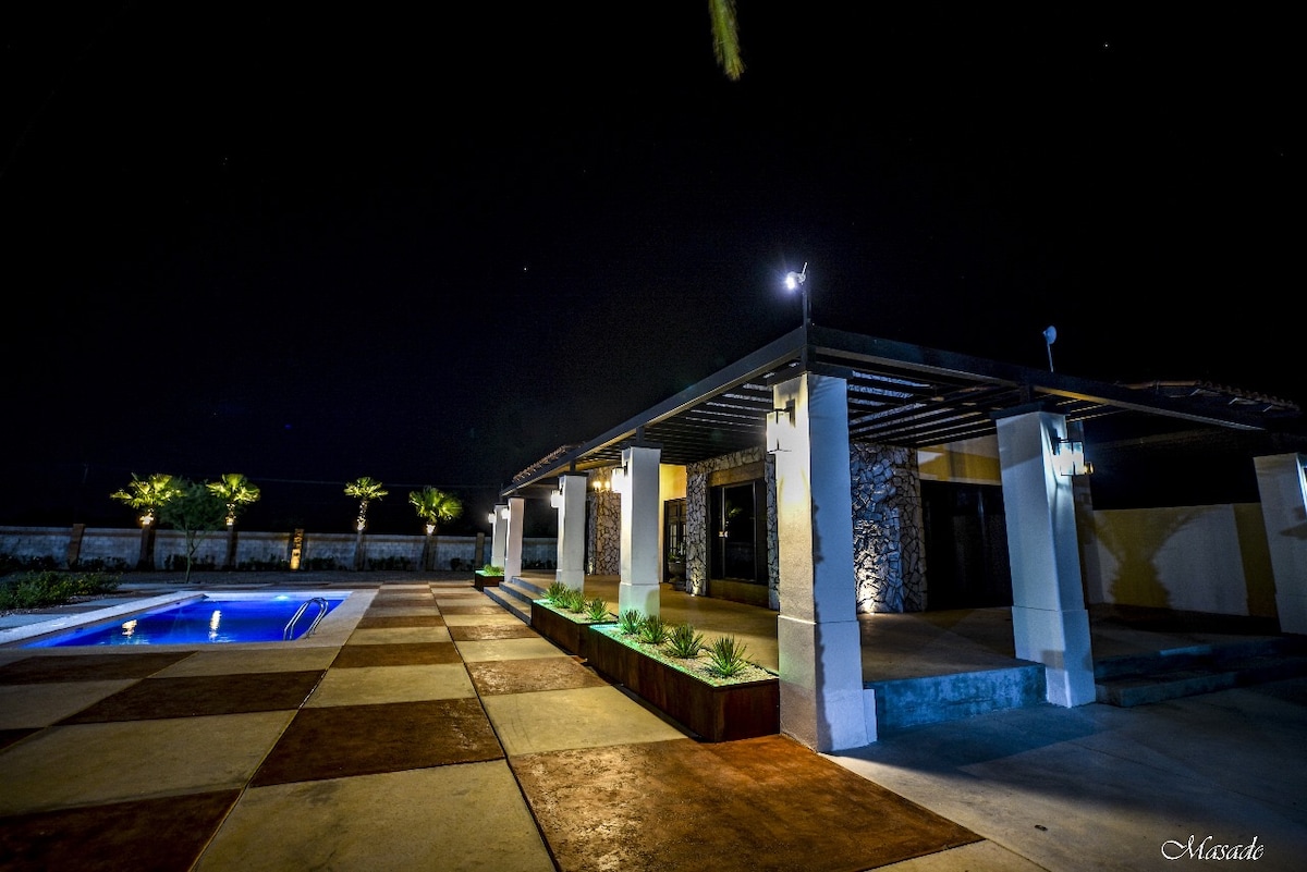 The exterior of the property is showcased at night, highlighting a calming pool area with subtle lights and well-maintained landscaping. The architecture features a blend of modern and rustic elements, with prominent use of stone and a spacious patio that connects to the surroundings.