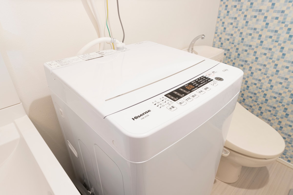 A modern washing machine is positioned next to a toilet, featuring a digital control panel with various settings. The space is brightened by light-colored walls, and a decorative blue mosaic tile adds visual interest in the background.