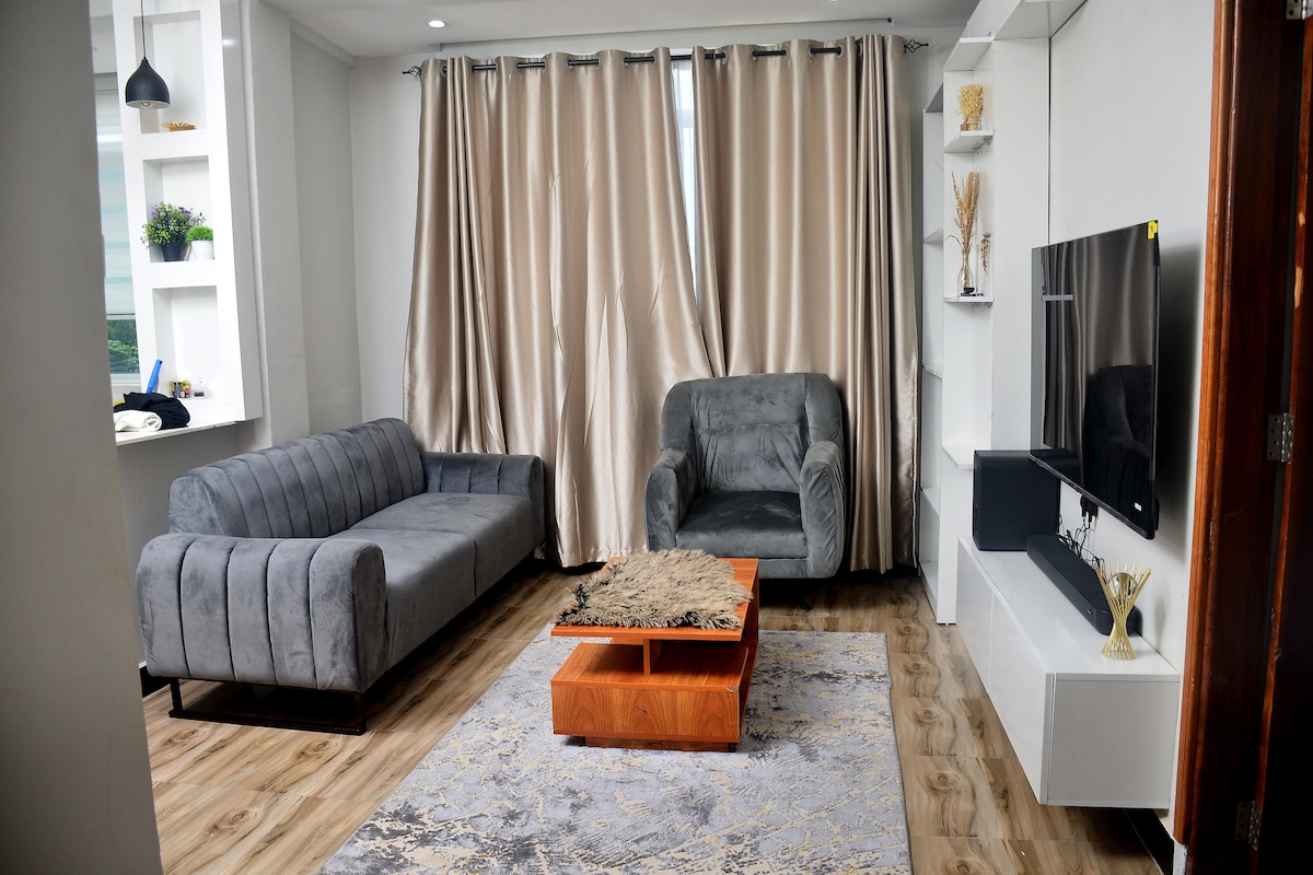 A comfortable sitting area features a gray sofa and an armchair with soft upholstery. A wooden coffee table is centered on a patterned rug, while large curtains frame a window, allowing for natural light. A mounted television is positioned on the wall opposite the seating.