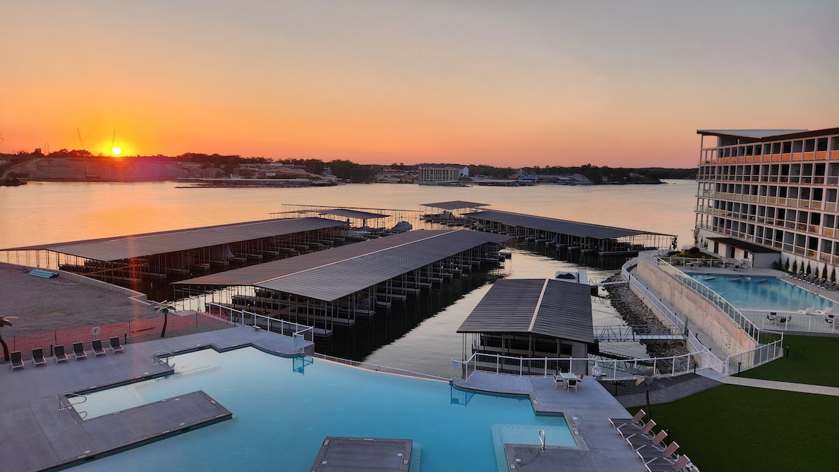 An impressive view showcases a serene sunset over the lake, reflecting vibrant hues across the water's surface. In the foreground, an expansive pool area is outlined by lounge chairs, while boat docks extend into the calm waters, framing the tranquil scene.
