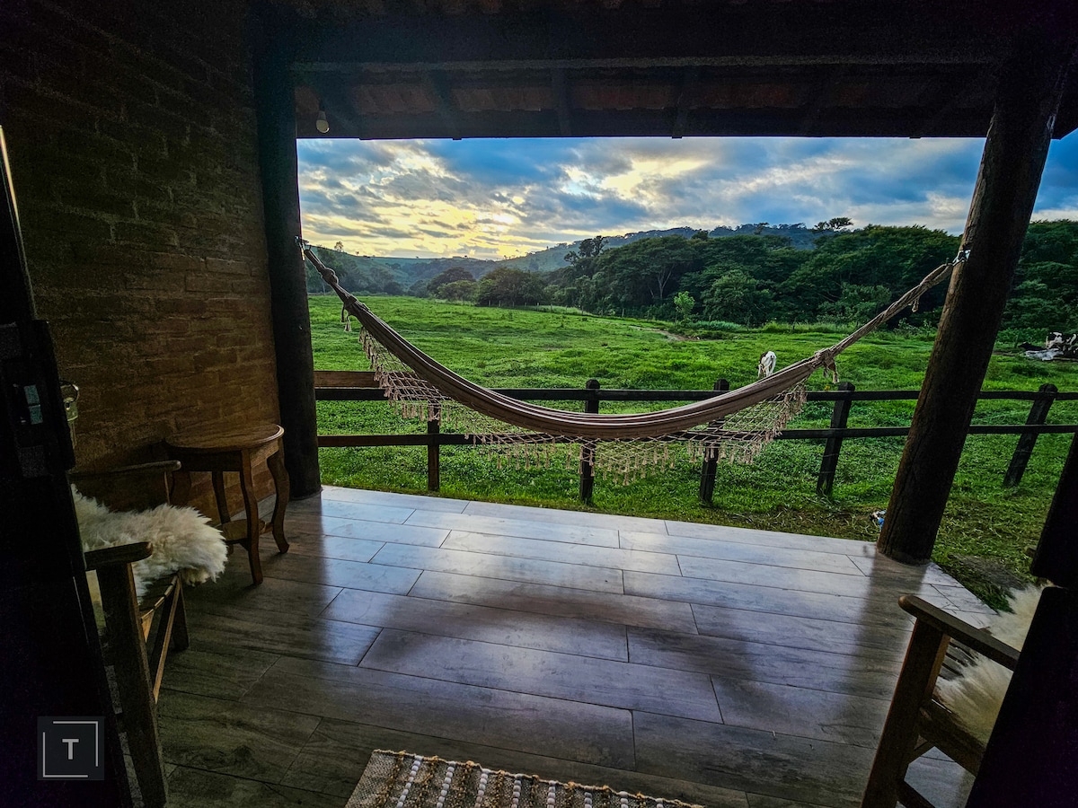 A spacious porch offers a view of lush green fields under a cloudy sky, featuring a hammock invitingly draped between wooden posts. Two rustic chairs are positioned nearby, contributing to a serene outdoor setting.
