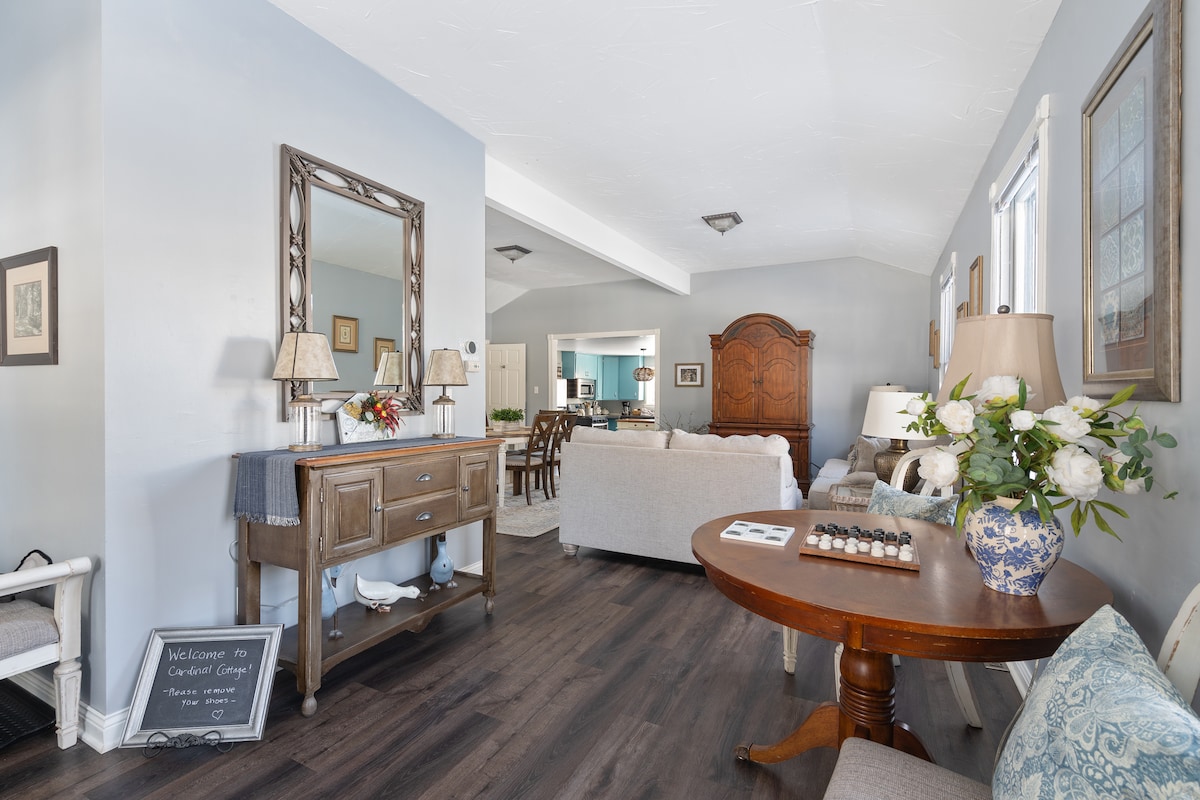 A spacious living area is highlighted by high ceilings and abundant natural light. A round table sits next to a welcoming arrangement of flowers, while a cozy seating setup invites relaxation. Decorative touches, including framed artwork and a mirror, enhance the room's charm.