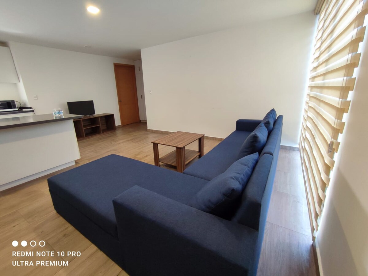 A modern living area is filled with natural light from a large window adorned with horizontal blinds. A dark blue sectional couch is positioned next to a wooden coffee table. A flat-screen television is placed on a low console across the room, enhancing the inviting space.