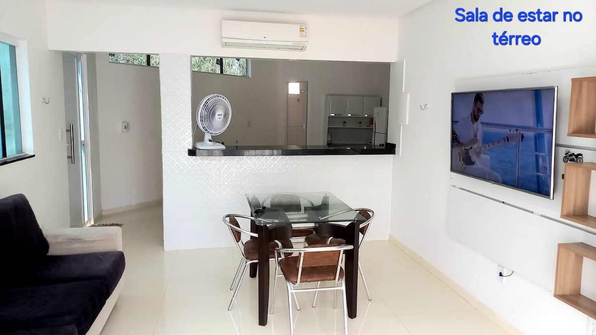 A modern living space features a glass dining table surrounded by four chairs. A comfortable sofa is positioned nearby. A wall-mounted television is visible, along with a fan for air circulation. Natural light floods the area through large windows.