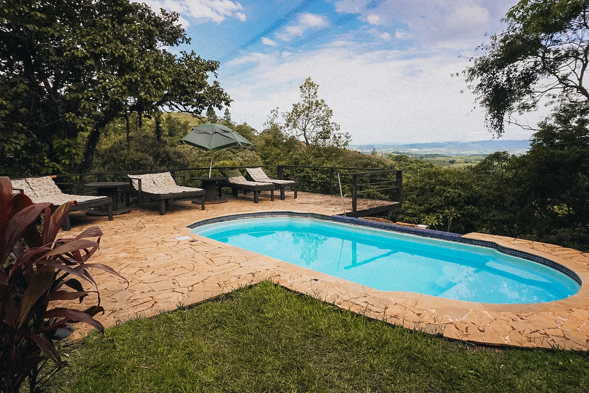 A natural setting features a swimming pool surrounded by lounge chairs. An umbrella provides shade, while lush greenery frames the area. The landscape offers expansive views of the mountains and valleys in the distance, creating a serene outdoor space for relaxation.