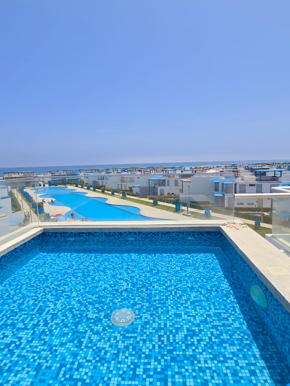 A private pool is displayed with vibrant blue tiles, set on a terrace overlooking expansive views of the ocean and surrounding properties. The infinity edge creates a seamless transition to the horizon, while the inviting water reflects the clear blue sky.