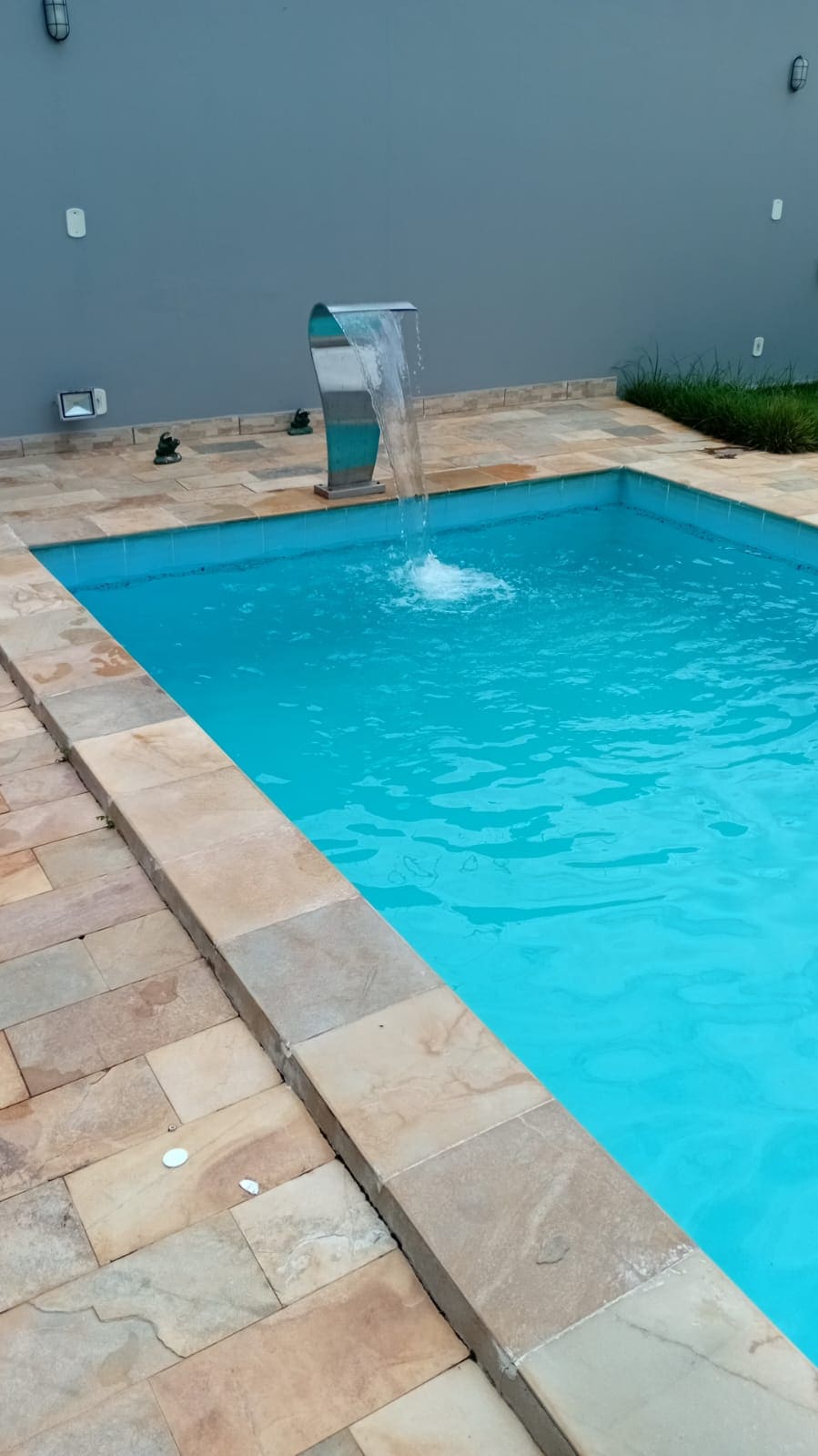 A serene swimming pool with bright blue water is featured, complemented by a waterfall feature. The pool is surrounded by a tiled patio and a grassy area, providing a relaxing outdoor space.