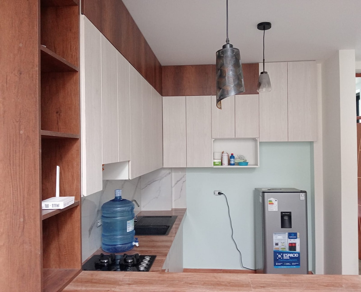 A modern kitchen displays sleek cabinetry in light and dark wood tones. A gas stove and refrigerator are present, along with a water dispenser. Adjustable pendant lighting hangs above, enhancing the functional space.