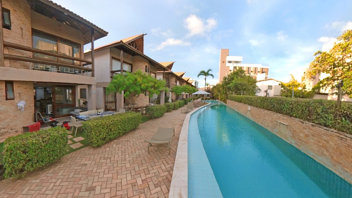 The outdoor area features a meandering pool surrounded by lush greenery and lounge chairs. Pathways lead between the residential buildings, offering access to various seating areas and umbrellas. A tranquil atmosphere is complemented by the presence of palm trees, enhancing the tropical environment.