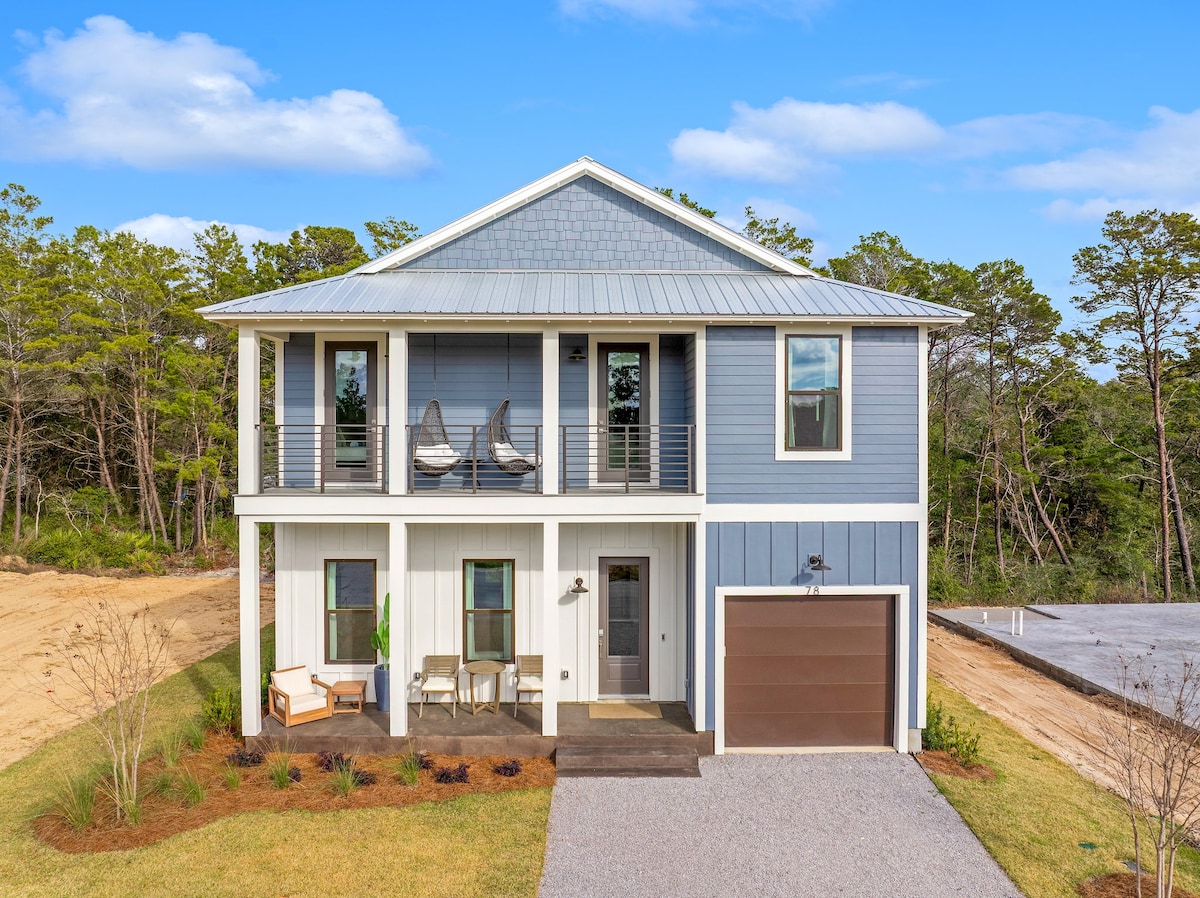 The exterior of a modern two-story home is shown, featuring a combination of blue and white siding. A covered porch with two hanging chairs and a small seating area invites relaxation. Fresh landscaping surrounds the home, with a gravel driveway leading to a garage on the right.