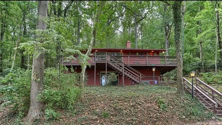 A two-story red wooden house is situated among lush greenery, surrounded by tall trees. A staircase leads up to a wrap-around deck, offering a view of the forest. The natural setting enhances the secluded feeling of the home.