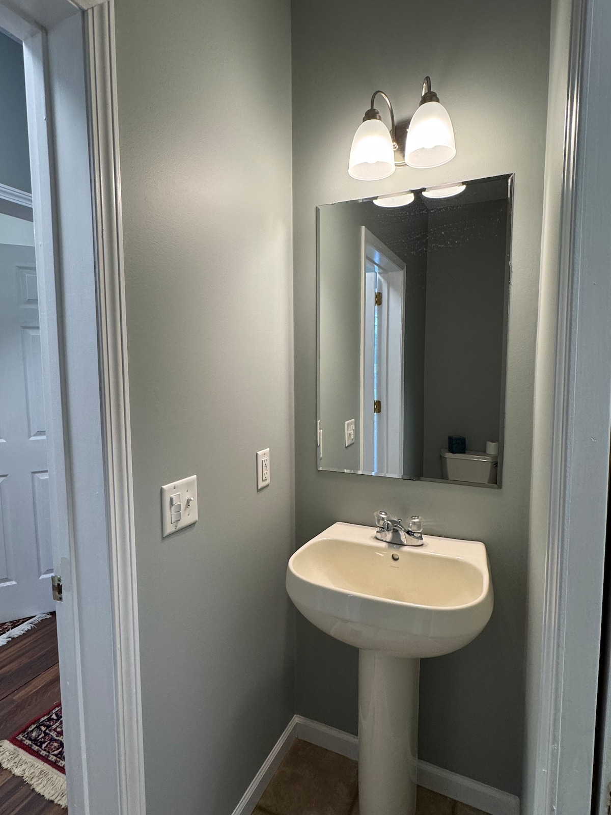 A small bathroom is presented, featuring a pedestal sink with dual faucet controls. A large mirror above reflects the space. Soft light is provided by two sconces mounted on the wall. The color palette includes calming light green walls and neutral flooring.