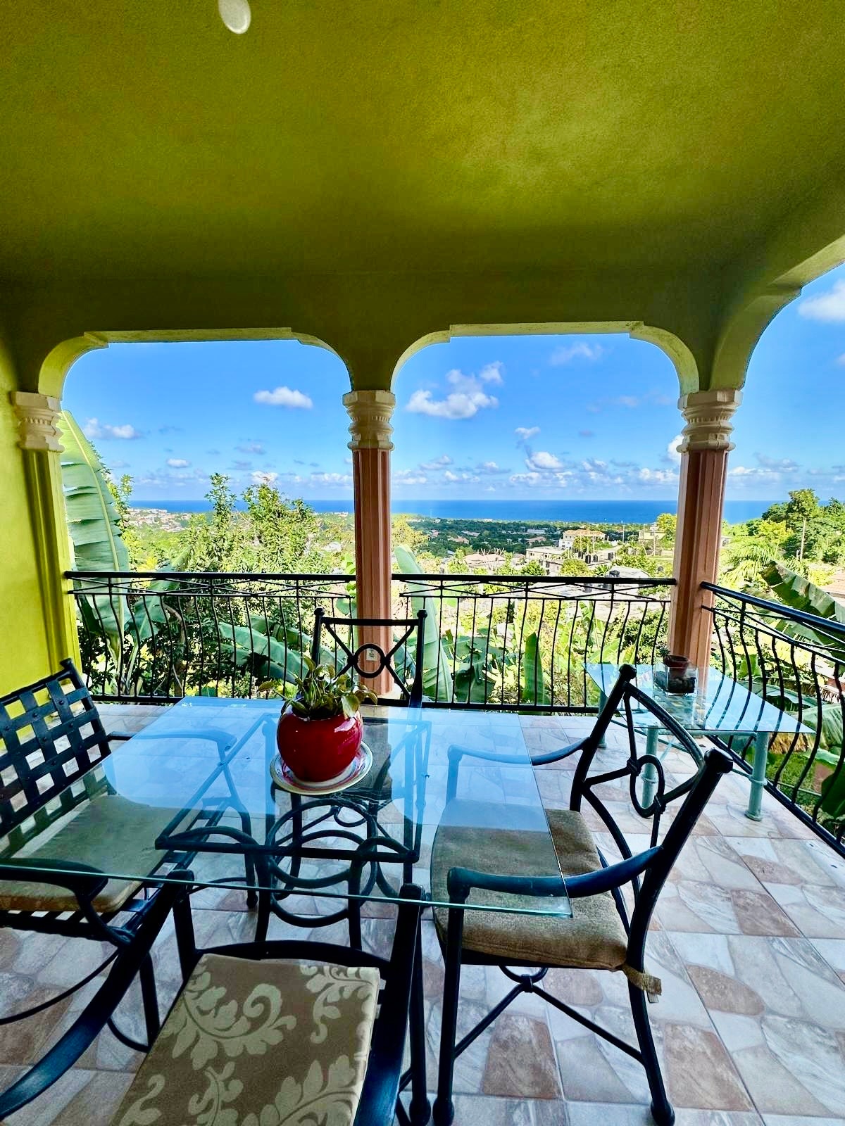 A dining area features a glass table surrounded by metal chairs, positioned in front of arched columns. The view reveals a scenic landscape with greenery and the Caribbean Sea under a bright sky, enhancing the outdoor dining experience.