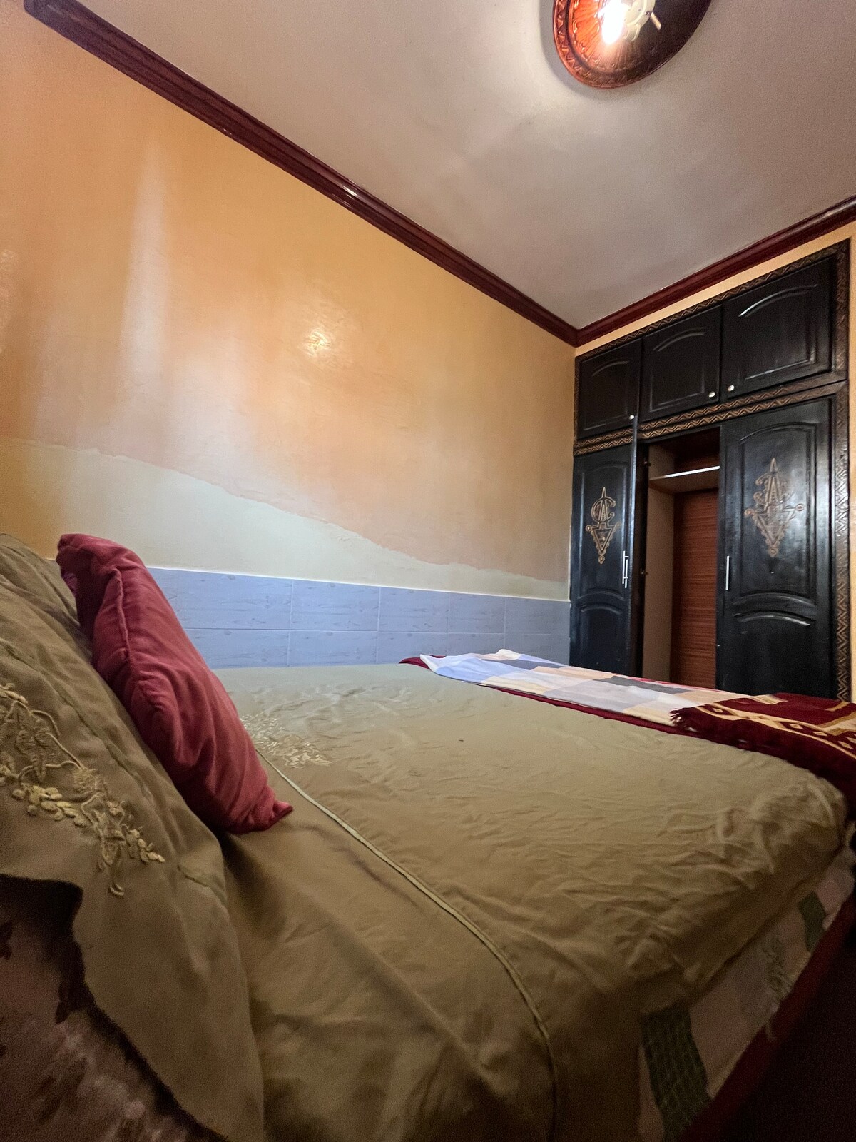 A cozy sleeping area features a spacious bed with an olive green coverlet and decorative pillows. The walls are painted in warm tones, complemented by a dark wood wardrobe with intricate designs. Soft light illuminates the space from a ceiling fixture.