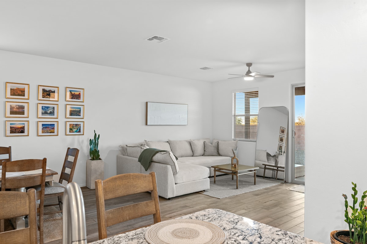 A light-filled living space features a spacious sectional sofa and a wooden dining table. Gallery-style wall art adds character, while a ceiling fan circulates air. Near the sofa, a full-length mirror is positioned, and sliding glass doors lead to a private outdoor area, enhancing natural light.