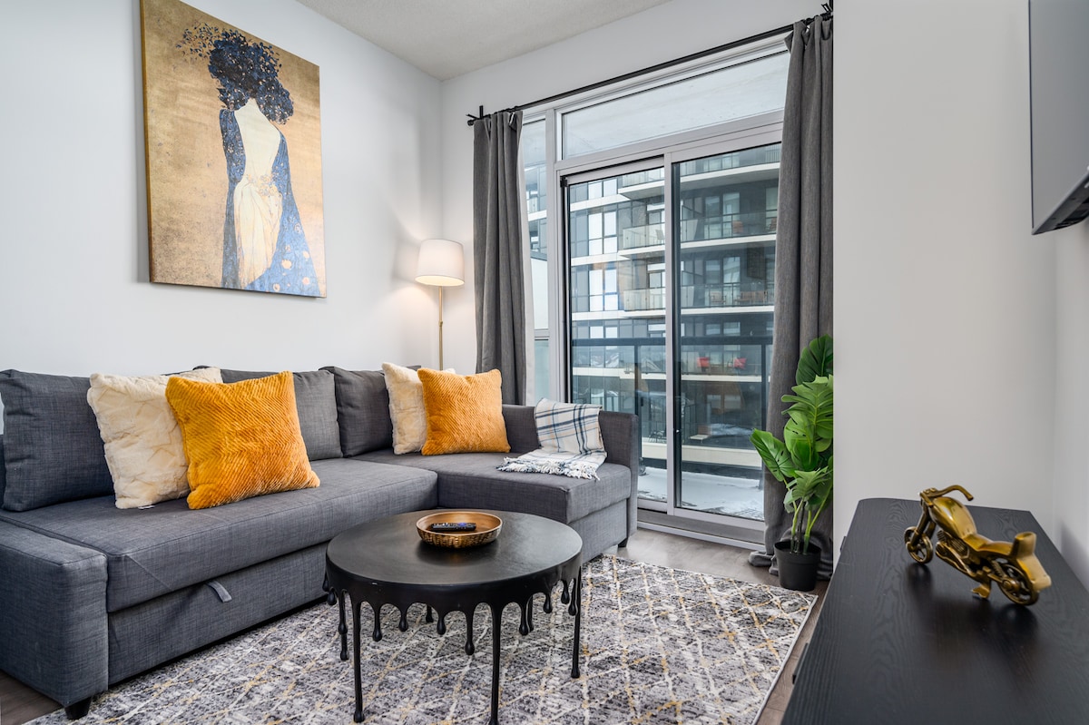 A contemporary living area is furnished with a dark sectional sofa, accented by orange and white cushions. A round black coffee table sits in the center, complemented by a patterned area rug. Large glass doors provide natural light and a view of the balcony.