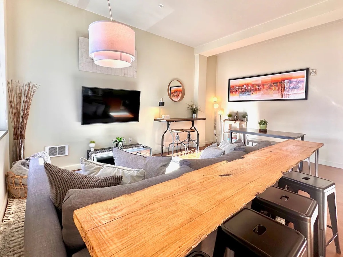 A spacious living area features a long wooden table accompanied by metal stools, providing ample seating. A comfortable gray sectional sofa is positioned near a wall-mounted television, while a circular light fixture hangs from the ceiling. Natural light enters through large windows, illuminating the modern decor.