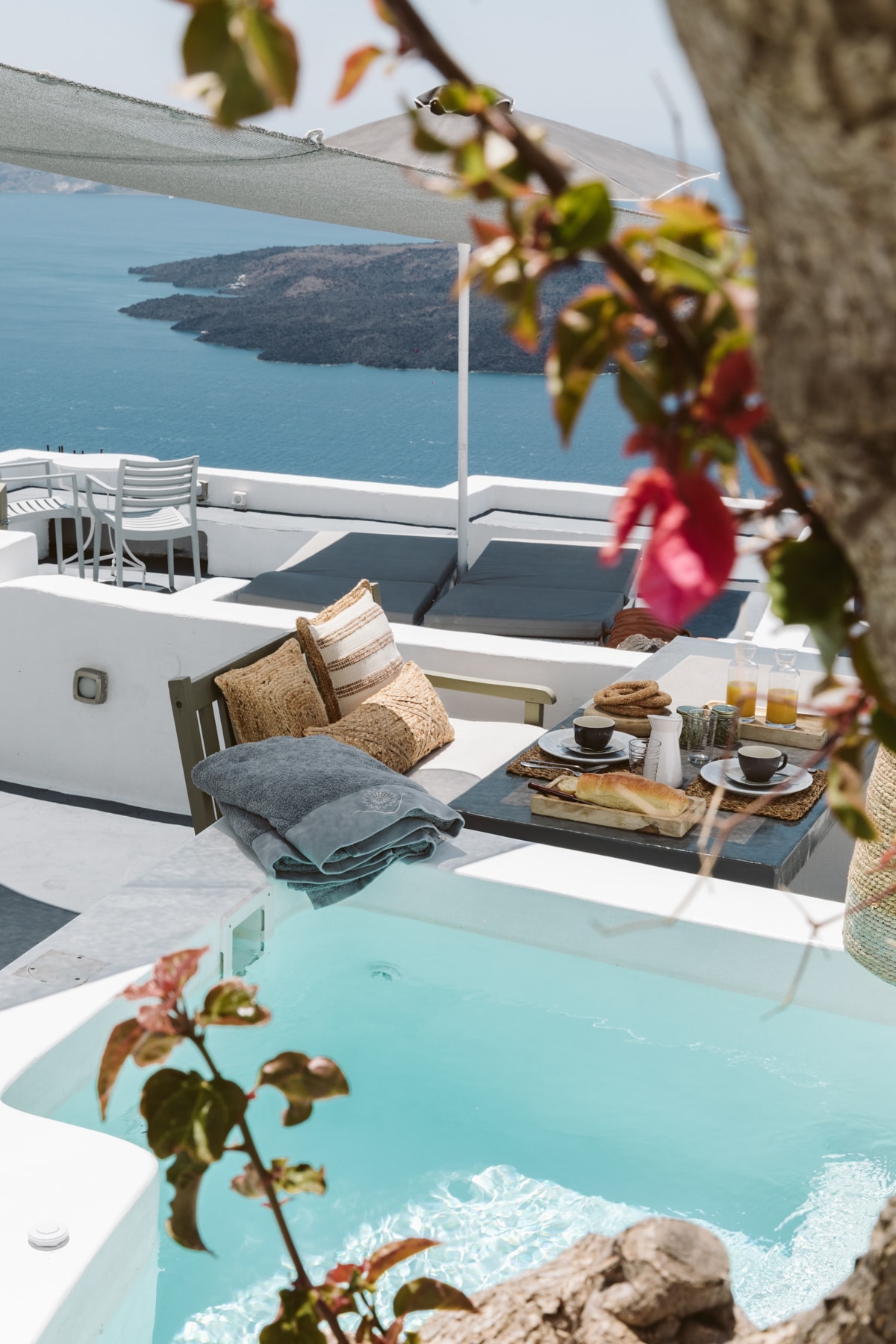 A serene outdoor space features a small Jacuzzi overlooking the Aegean Sea. A table, set for breakfast, is adorned with a selection of food and drinks. Comfortable seating, surrounded by vibrant flowers, enhances the peaceful atmosphere of the area.