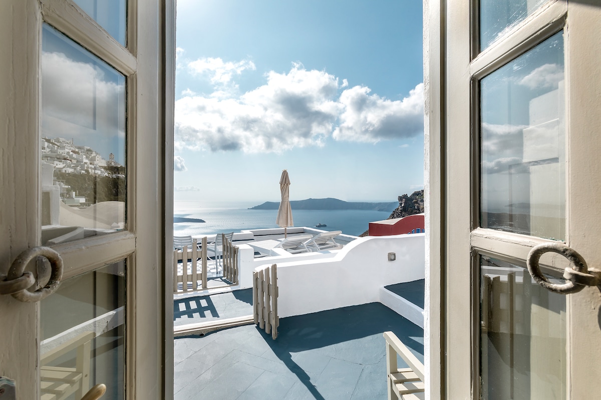 Double doors open to reveal a panoramic view of the Aegean Sea. The scene captures sun loungers arranged on a sunlit terrace, with a large umbrella standing in the center, set against a backdrop of distant cliffs and cloudy blue skies.