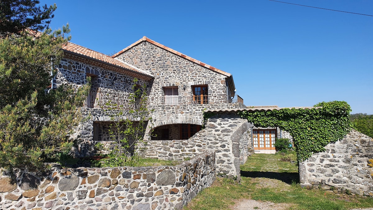 A spacious stone house is depicted, featuring a rustic facade with a slate roof and wooden windows. Lush greenery surrounds the entrance, while a path leads through a landscaped area. The clear blue sky above enhances the serene outdoor environment.