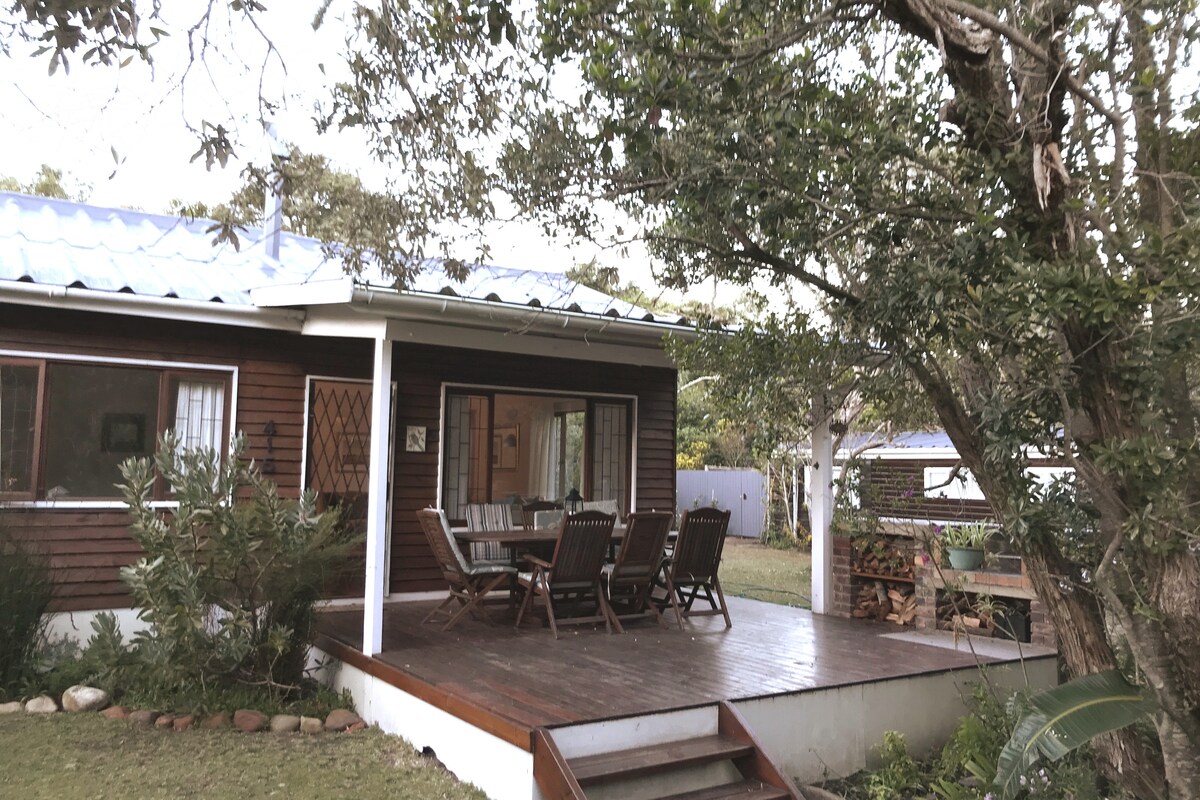 A wooden deck extends from the main house, featuring a set of outdoor dining furniture. Surrounding trees provide shade, while a small staircase leads to the yard. The entrance to the home is visible, framed by large windows, giving a glimpse of the interior.