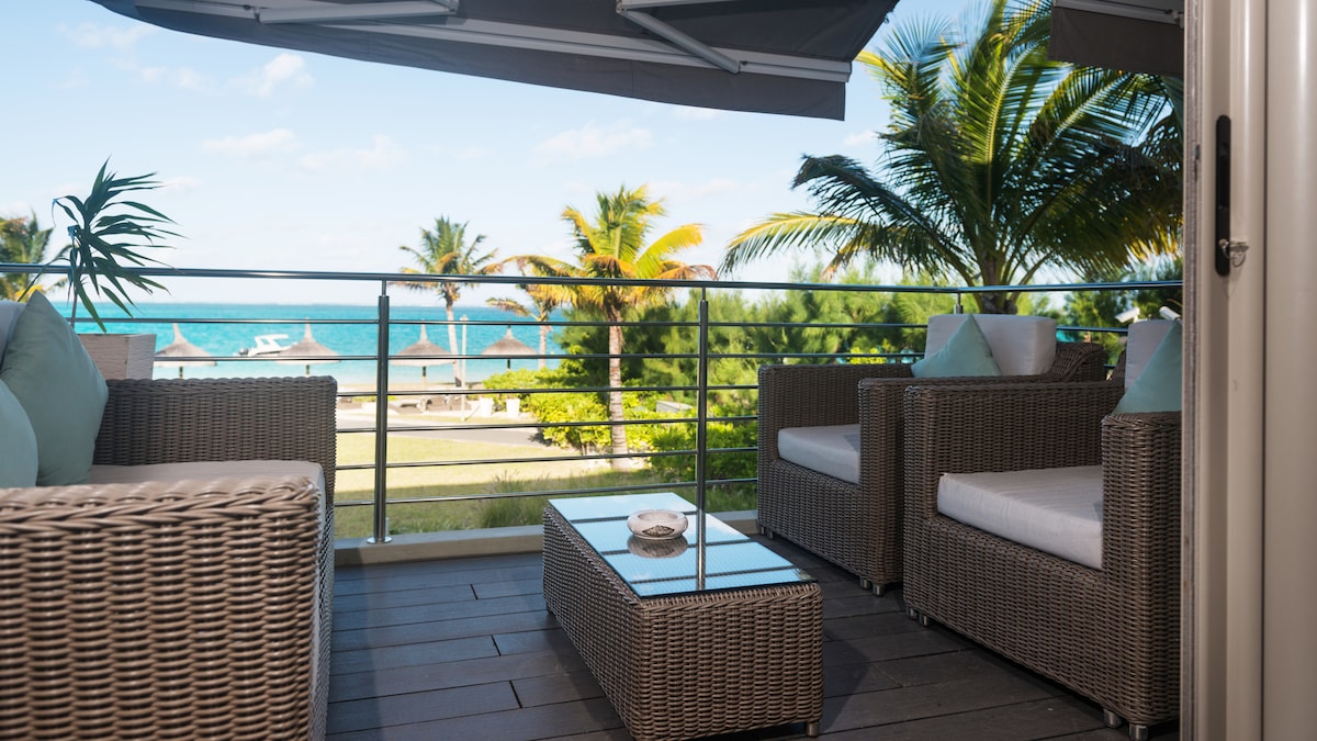 A private terrace is featured with comfortable wicker seating and a glass-topped coffee table. Lush green foliage and palm trees surround the area, with views of the turquoise lagoon and distant beach umbrellas creating a serene outdoor space.