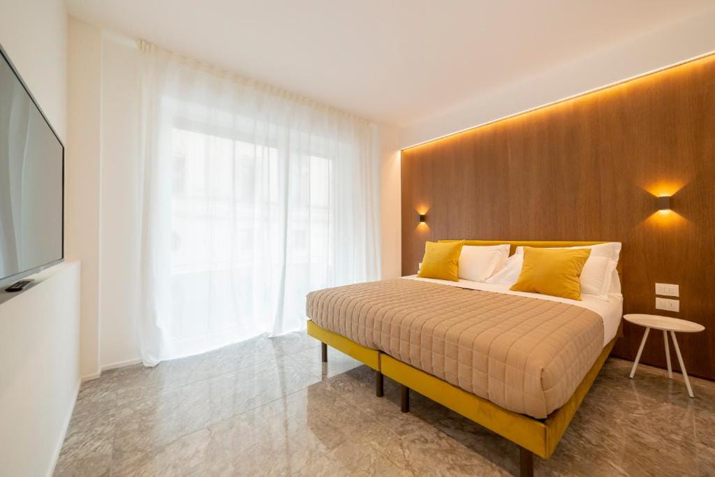 A spacious bedroom features a large bed with yellow accent pillows, paired with a minimalist side table. Natural light filters through sheer white curtains, highlighting the smooth, marble flooring and warm wooden wall accents.