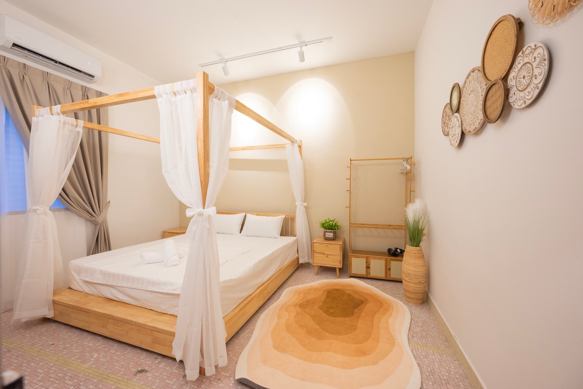 A serene sleeping area features a wooden four-poster bed with a clean white mattress and bedding. A natural fiber rug adds warmth to the space. Light walls provide a calming backdrop, complemented by decorative wall hangings and soft curtains framing a window.