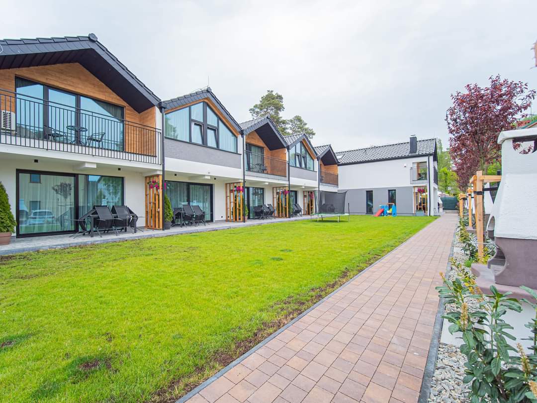 A spacious outdoor area, featuring a manicured lawn and a pathway made of paving stones. Modern structures with pitched roofs are visible, each equipped with balconies. Black lounge chairs are arranged on the patios, offering a comfortable seating option for guests.