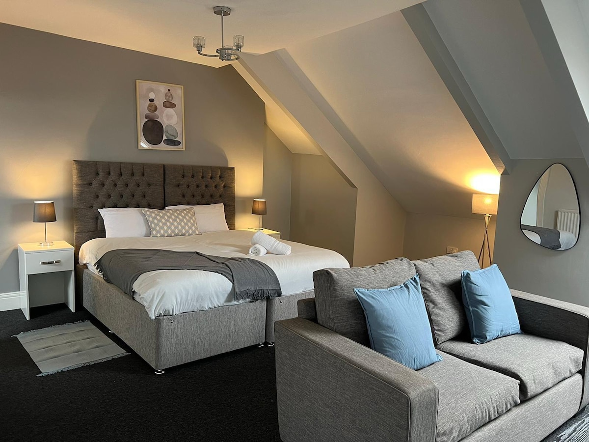 A spacious bedroom features a tufted headboard bed with soft linens and accent pillows. A cozy seating area is provided by a grey sofa adorned with blue cushions. Warm lighting is created by bedside lamps and a ceiling fixture, enhancing the room's inviting atmosphere.