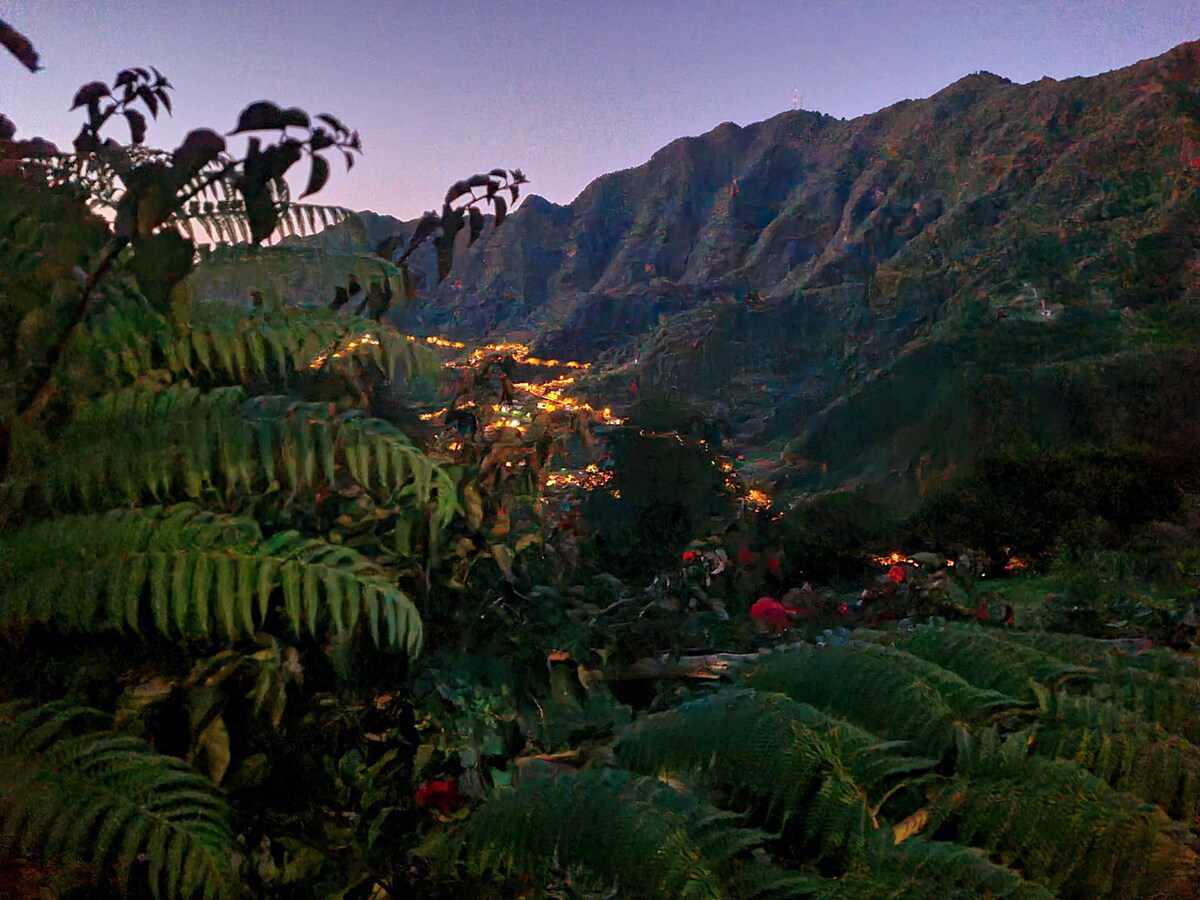 A serene mountainous landscape is depicted, featuring lush green foliage in the foreground. The hillside is illuminated by warm lights from a distant settlement, creating a gentle contrast against the darkening sky.