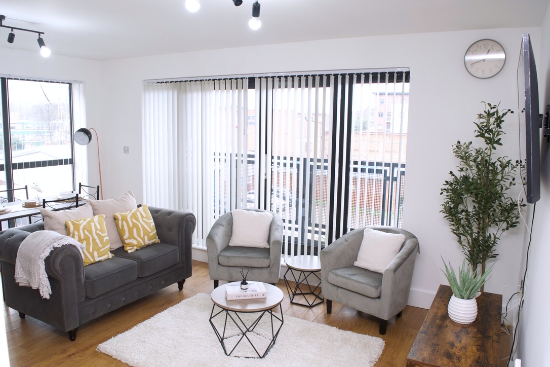 A modern living room features a grey sofa with patterned cushions and two armchairs. A round coffee table is centered on a plush rug, and a wall clock is mounted above. Large windows with vertical blinds allow natural light to fill the space.