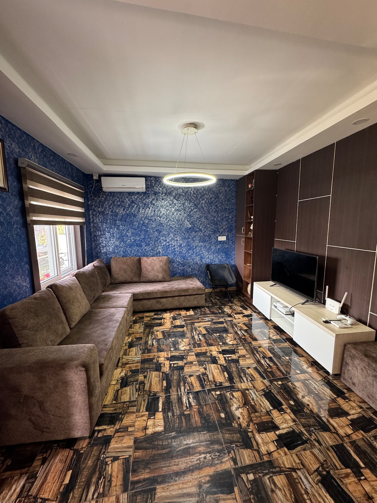 A modern living area is designed with a large brown sectional sofa, positioned against a textured blue wall. A circular light fixture hangs from the ceiling, illuminating the room. A sleek media console holds a television, while a window with striped shades allows natural light to enter.