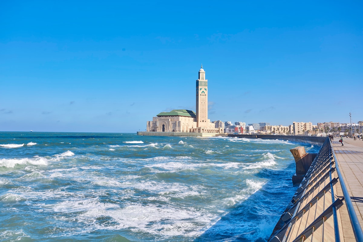 The image showcases the Hassan II Mosque rising prominently against the clear blue sky, located by the ocean. Waves crash against the shoreline, while a promenade lined with railings invites leisurely strolls along the coast.
