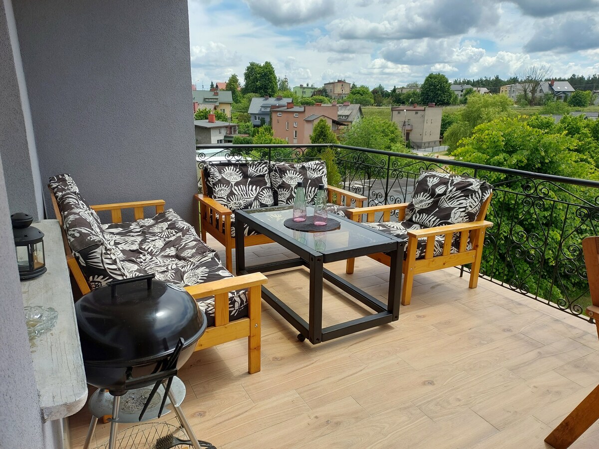 An outdoor balcony is furnished with comfortable seating featuring floral-patterned cushions and a glass-topped table. A small grill is positioned nearby, and a view of greenery and buildings is visible in the background under a partly cloudy sky.