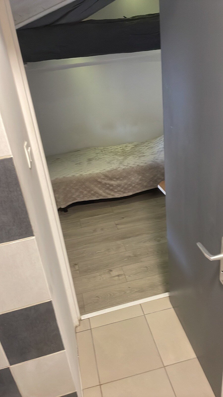 A view into a compact studio space is presented, featuring a neatly made bed with a light-coloured bedspread. The flooring is styled with wooden planks, and a doorway indicates the entrance to the room. Walls are minimally decorated, enhancing the sense of space.