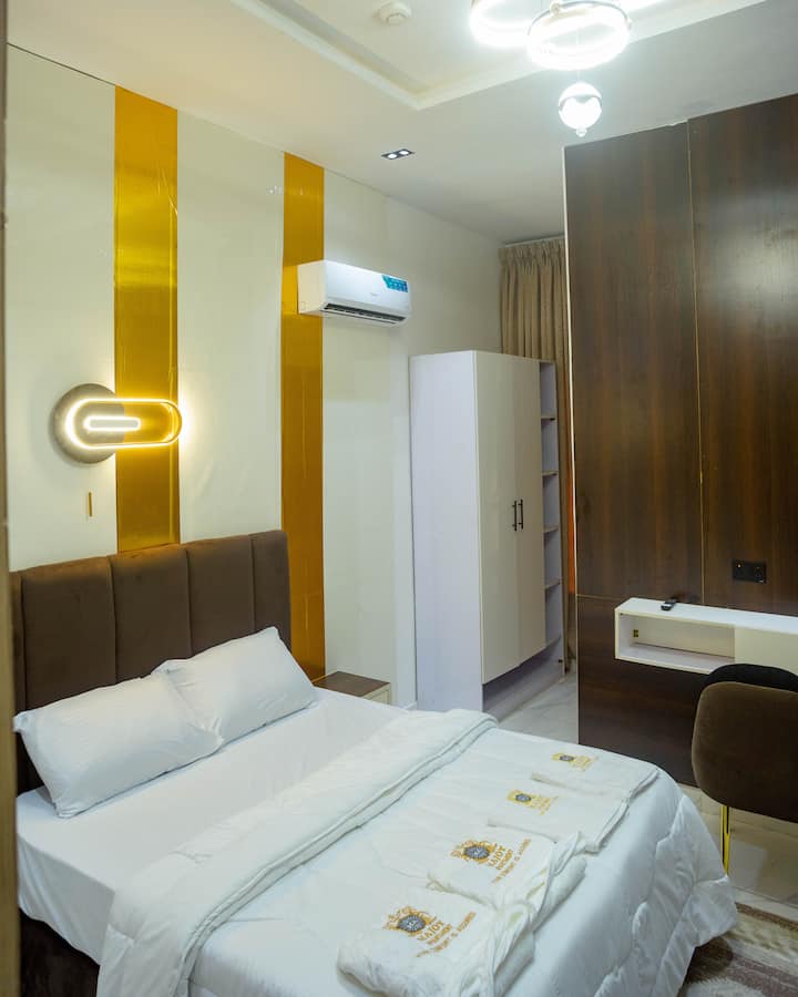 Un dormitorio elegante y acogedor con una lujosa cama doble, un armario blanco empotrado, aire acondicionado y un espacio de trabajo dedicado con un elegante escritorio y una silla. La iluminación suave y los detalles dorados crean un ambiente cálido y acogedor... perfecto para descansar o trabajar a distancia.