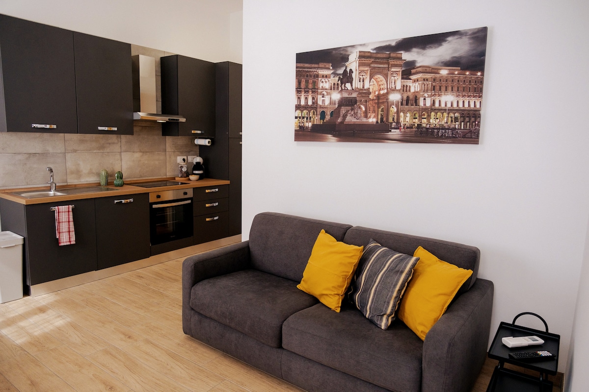 A modern kitchen area features dark cabinetry and stainless steel appliances, including an oven and coffee maker. A comfortable gray sofa, adorned with yellow and striped cushions, invites relaxation. A wall art piece depicting a landmark adds character to the space.