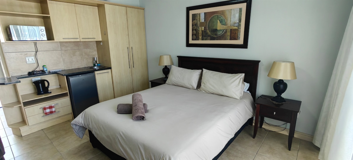 A spacious room features a queen-size bed with light bedding, complemented by two bedside lamps. A kitchenette area is visible, equipped with a refrigerator and a microwave. Artwork adorns the wall above the bed, and a pair of towels is neatly placed on the bed.